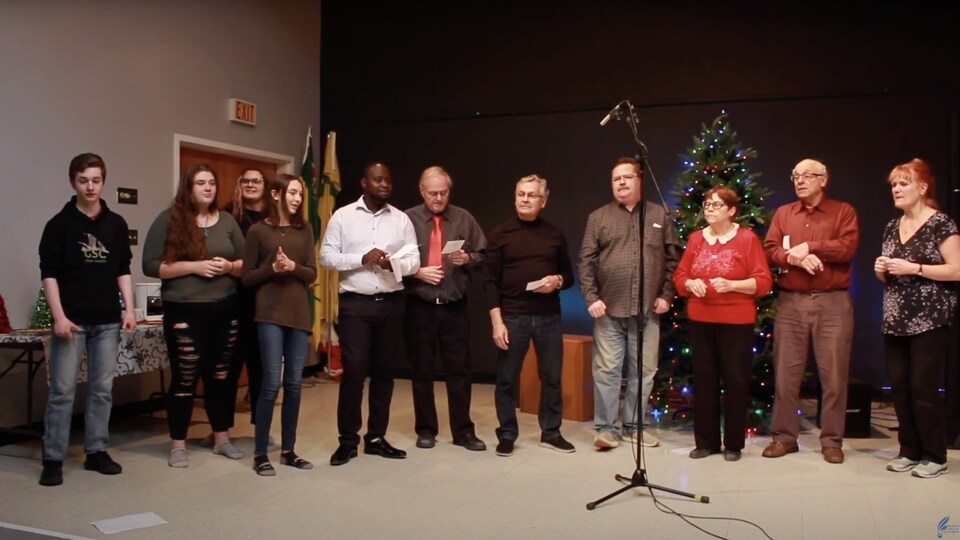 Un groupe intergénérationnel réuni pour chanter devant un microphone, un sapin de Noël est visible en arrière-plan.