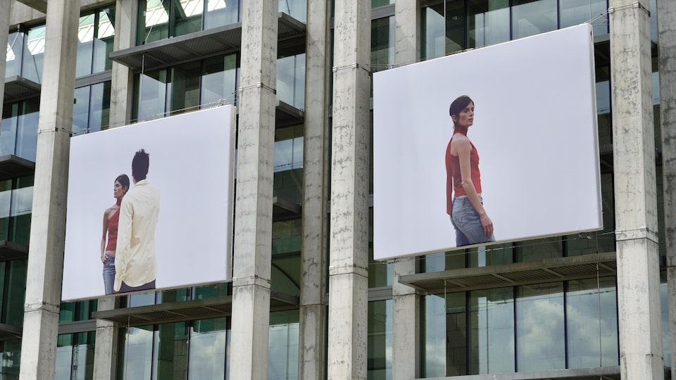 Plan rapproché de la façade du MBAC sur laquelle on voit un accrochage d'une série de photos sur fond blanc représentant un femme et un homme.