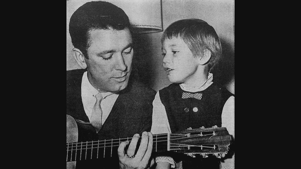 Le chanteur Georges La Flèche joue de la guitare à côté d'un enfant.