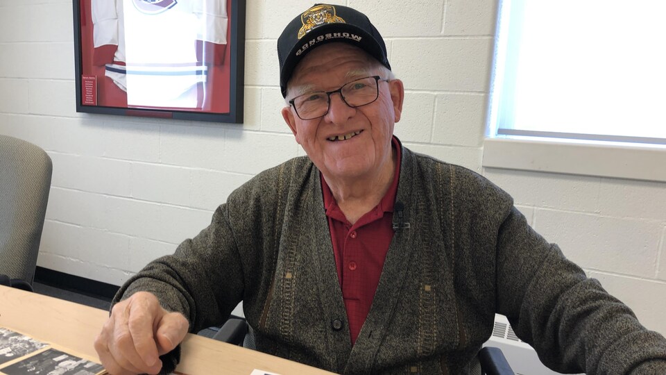 Homme souriant avec lunettes et casquette avec des photos sur la table devant lui.