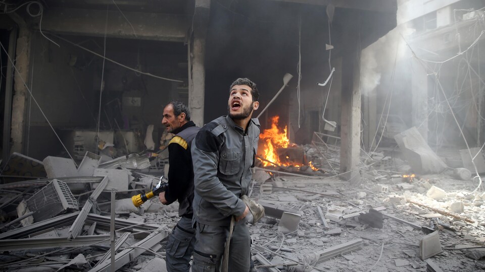 Un homme avec une hache regarde le ciel, alors qu'un autre tient une lance d'incendie, parmi les décombres.