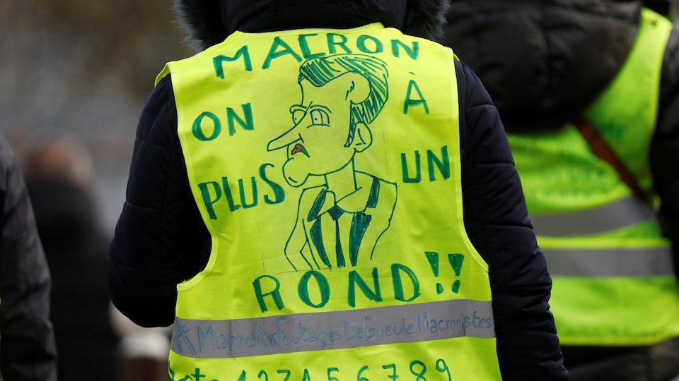 Un manifestant porte un gilet jaune dans le dos duquel il est écrit: « Macron on n'a plus un rond! » avec une caricature d'Emmanuel Macron.