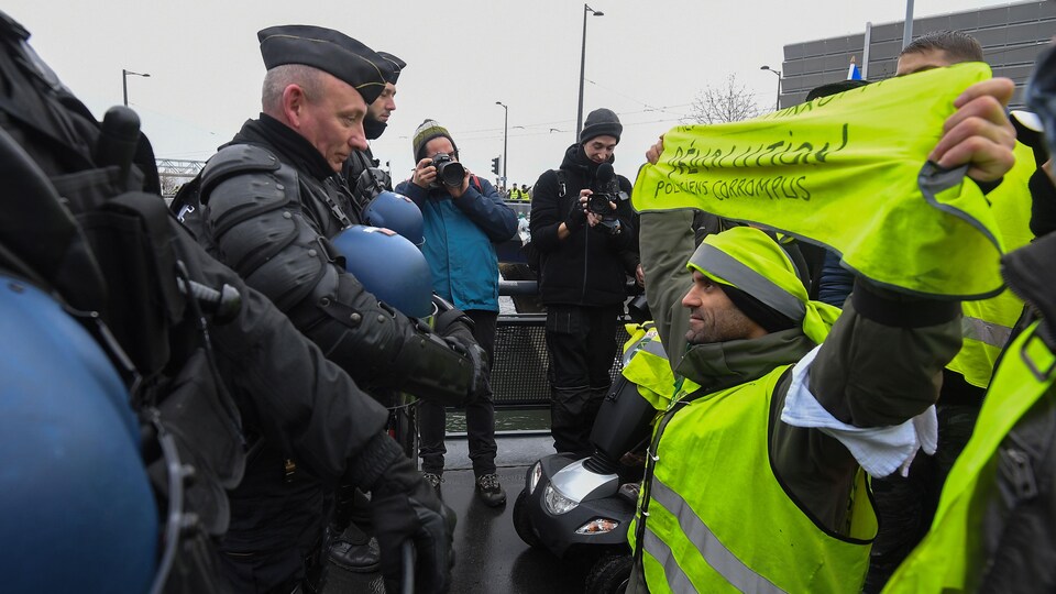 Un gilet jaune à genoux devant un policier brandit une pancarte où on peut lire  : « Révolution – Politiciens corrompus ».