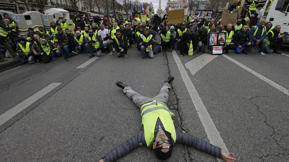 Un homme couché en pleine rue; des dizaines de gilets jaunes derrière, agenouillés. 