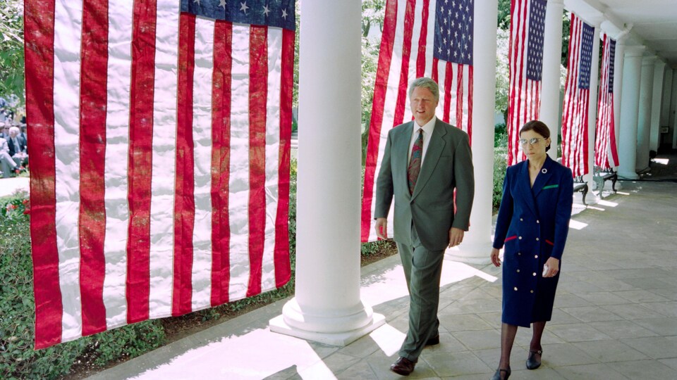 Bill Clinton et Ruth Bader Ginsburg marchent côte à côte devant des drapeaux américains.