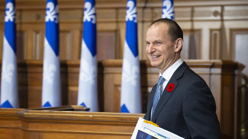 Plan moyen d'Eric Girard debout, tenant une copie de la mise à jour économique dans sa main gauche.