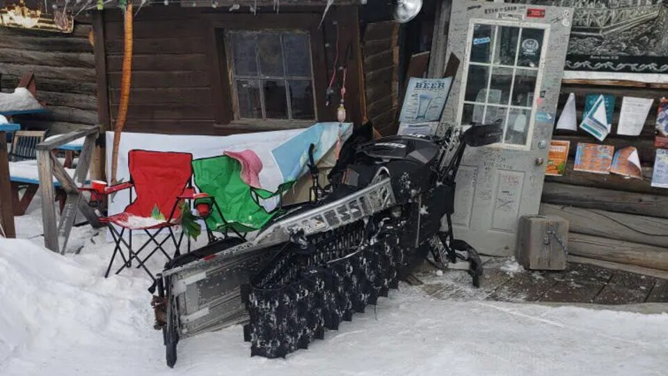 Une motoneige renversée près de chaises devant une bâtisse en bois rond.