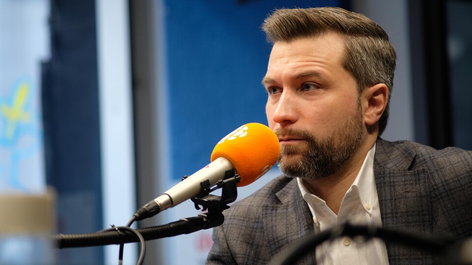 Un homme avec une courte barbe derrière un micro dans un studio de radio.