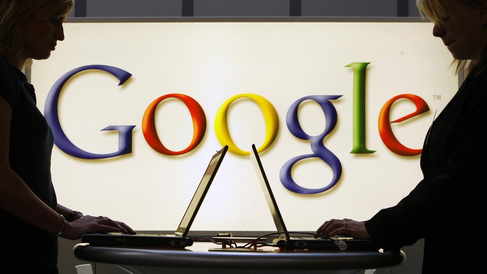 Deux femmes debout travaillent face à face sur des ordinateurs portables posés sur une table. En arrière-plan, le logo de Google est visible sur un écran lumineux.