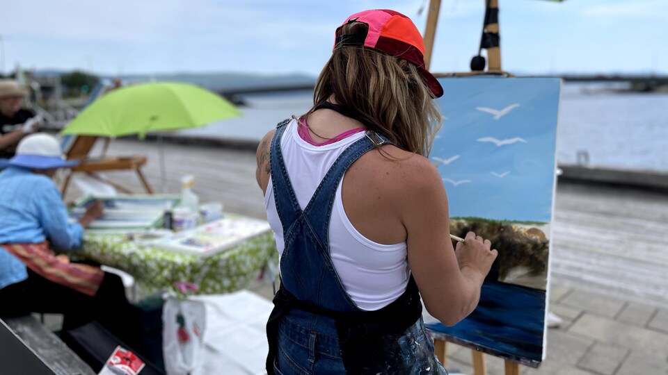 Une femme de dos peint un tableau du paysage marin devant elle.