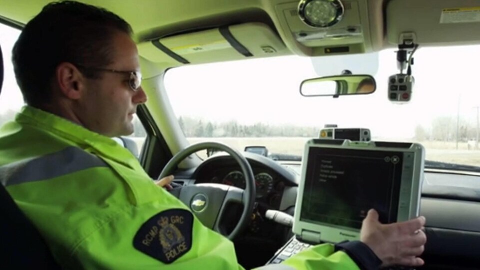 Un agent de la GRC porte un gillet vert. Il est derrière un volant dans une voiture.