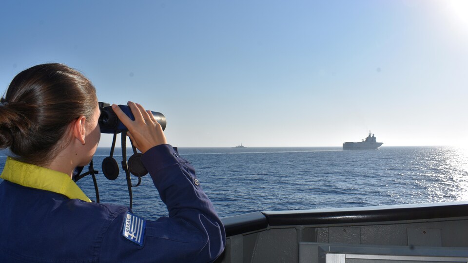 Une militaire sur un bateau en observe un autre, au loin, avec des jumelles.