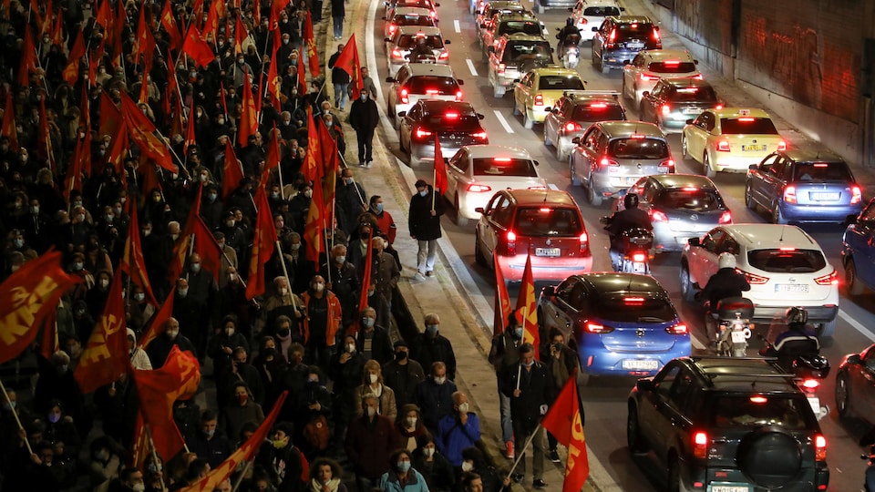 Des centaines de personnes brandissent un drapeau rouge à côté d'une avenue bloquée par des voitures.