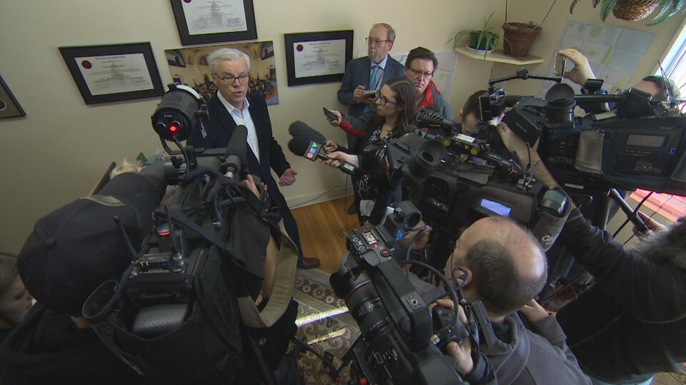 Greg Selinger, au fond, entouré de caméras, micros et journalistes dans son bureau de circonscription