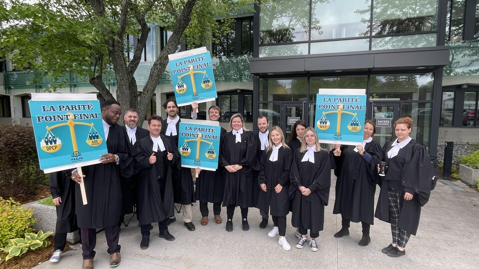 Une dizaine d'avocats portent une toge devant le palais de justice de Val-d'Or. Ils brandissent des pancartes demandant la parité.
