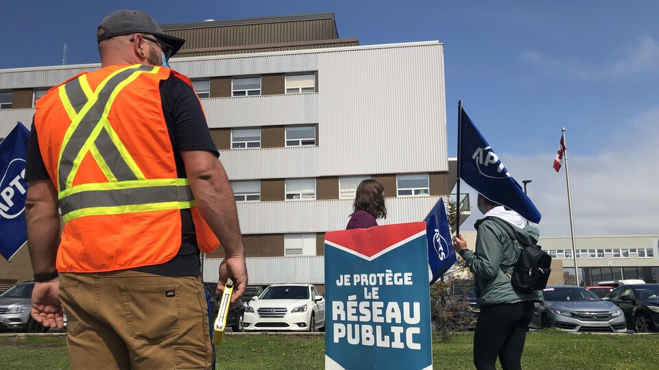 Trois travailleurs syndiqués avec l'APTS ont des drapeaux aux couleurs du syndicat dans les mains et font du piquetage devant l'hôpital de Sept-Îles.