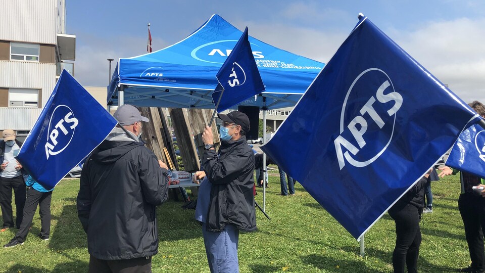 Cinq travailleurs syndiqués avec l'APTS ont des drapeaux aux couleurs du syndicat dans les mains et sont devant un chapiteau érigé pour l'occasion.