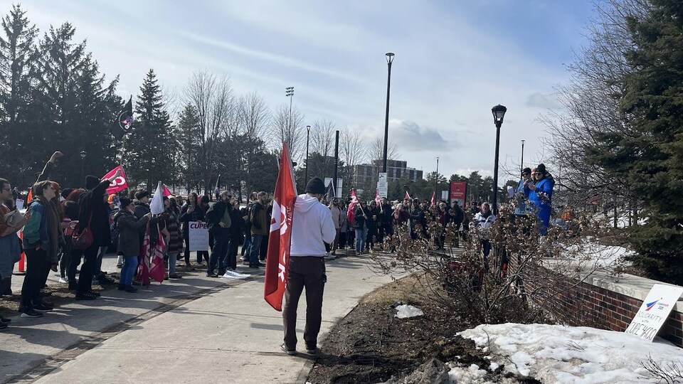 Plusieurs personnes sont debout et en écoutent une autre parler. On aperçoit le drapeau du syndicat et quelques pancartes dans la foule. 