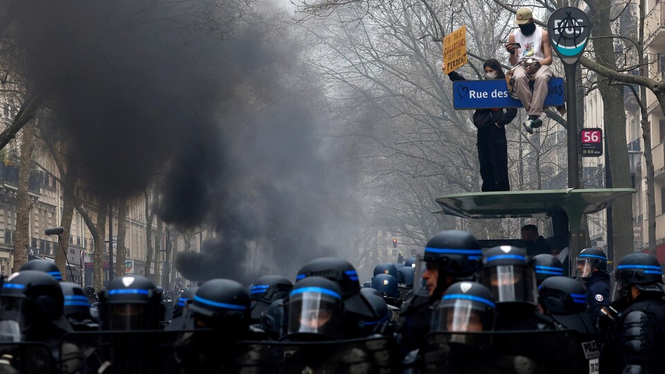 Des manifestants se tiennent sur une structure surélevée alors que des policiers casqués font bloc dans la rue, dans un ciel enfumé.
