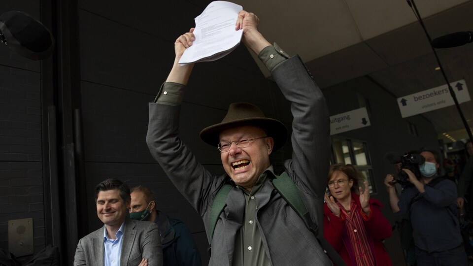 Un homme brandit un document avec un air de victoire, entouré de personnes qui applaudissent.