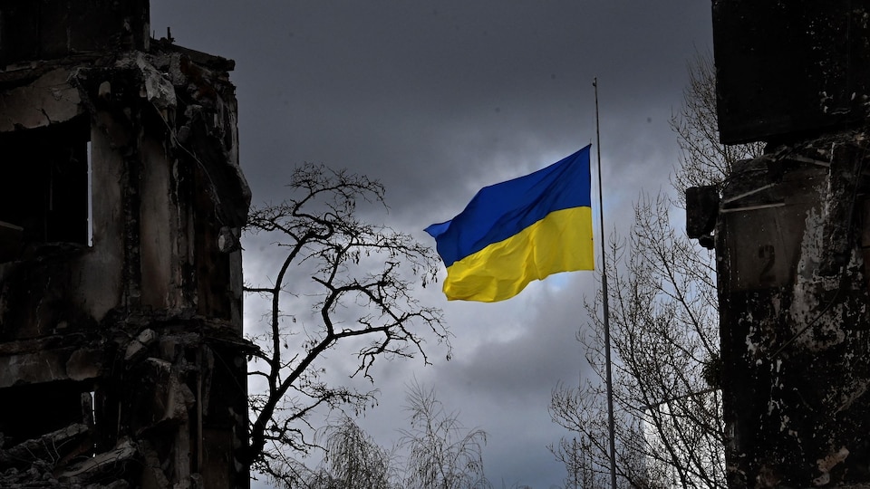 Un drapeau ukrainien flotte entre des bâtiments détruits par un bombardement.