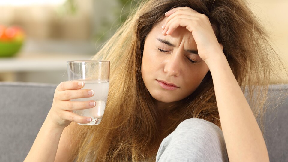 Une jeune femme, les yeux fermés, se tient la tête d'une main avec un verre d'eau dans l'autre.