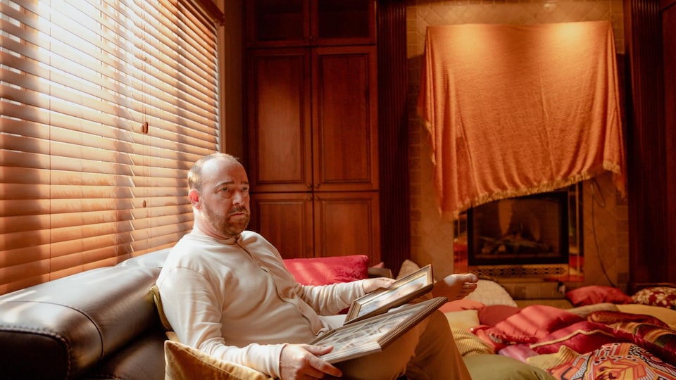 The actor is sitting on a couch and holding frames in an orange living room. 