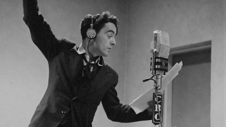 Dans un studio de radio, derrière un microphone sur pied, le réalisateur Guy Mauffette les bras en l'air.