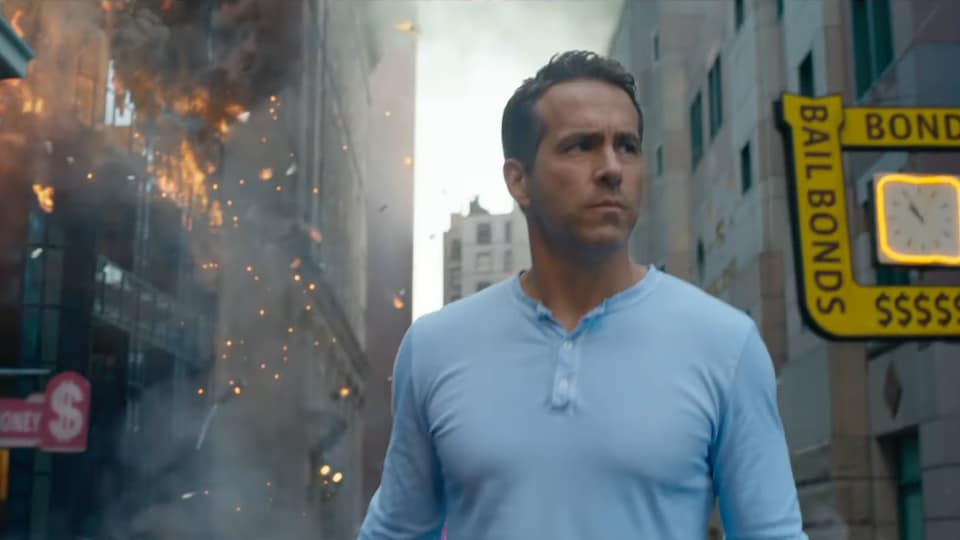 Un homme se tient au milieu d'une rue dans une ville. Des explosions ont lieu derrière lui dans des bâtiments de plusieurs étages.