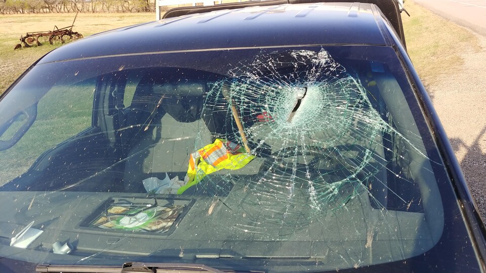 Vue de face d'un pare-brise brisé avec un trou longitudinal au niveau de la tête du conducteur. Un mache de hache est entre les sièges du conducteur et du passager.