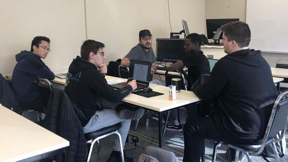 Cinq étudiants discutent autour d'une table dans une salle de classe. Des ordinateurs sont ouverts devant eux.