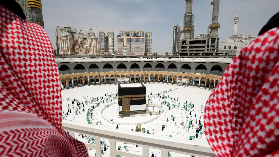 Deux hommes sur un balcon contemple des dizaines de personnes qui marchent en cercle autour de la Kaaba, une grande structure cubique au centre d'une esplanade.