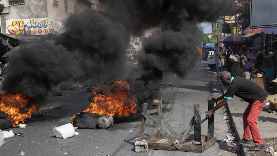 Un homme bloque une rue de Port-au-Prince en Haïti, alors que des pneus brûlent et dégagent une dense fumée noire.