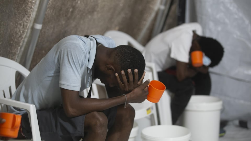 Deux hommes assis sur des chaises sont penchés par en avant au-dessus de seaux et tiennent des tasses.