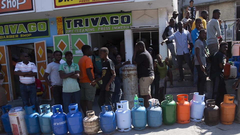 Une vingtaine de personnes, principalement des hommes, font la file devant des magasins avec leurs bidons de propane dans une rue commerciale de Port-au-Prince.