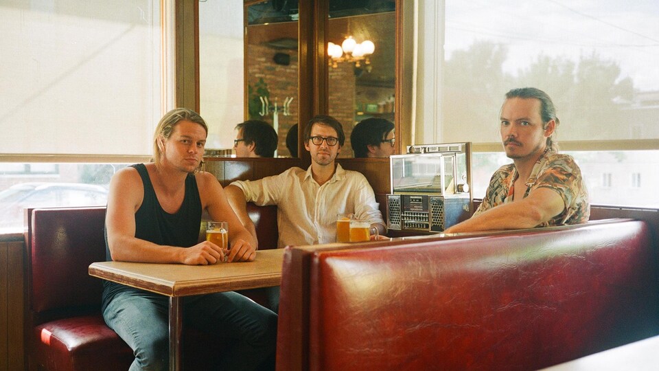 Trois jeunes hommes sont attablés à une banquette de restaurant, près d'un juke-box.