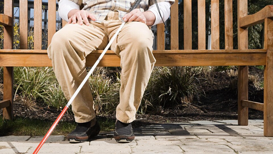 Un homme handicapé visuel assis sur un banc avec sa canne blanche.