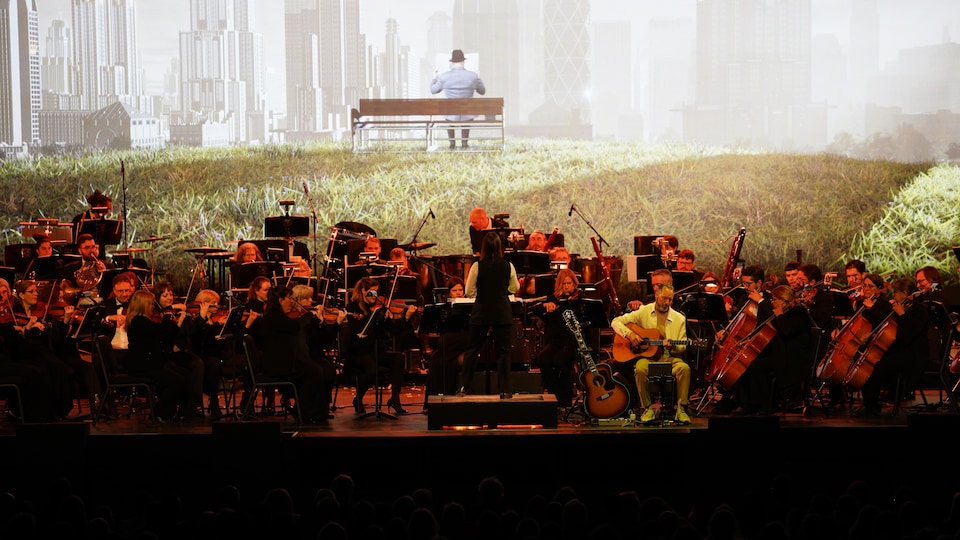Un orchestre symphonique est sur scène; Derrière lui, la projection de l'image d'un homme sur un banc de parc.