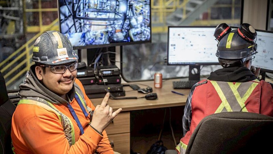 Deux employés de la mine portant des habits de constructions, assis devant un ordinateur. Un des deux employés est tourné vers la caméra, il sourit et fait un geste avec ses mains.