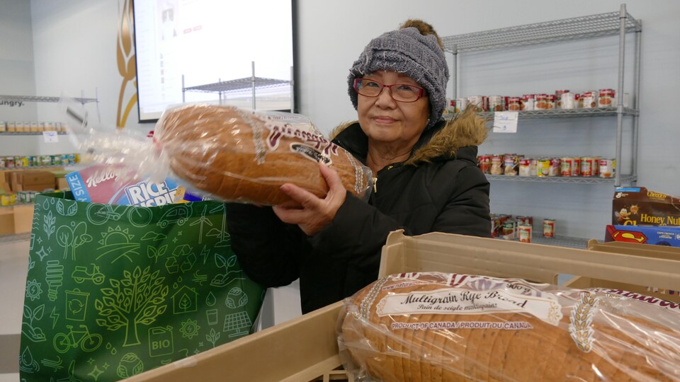 Erlinda De Guzman tientu pain dans ses mains, à côté de son sac rempli de denrées. Elle est dans le local de Harvest Manitoba et on peut voir autour d'elle des tablettes remplis de produits. 