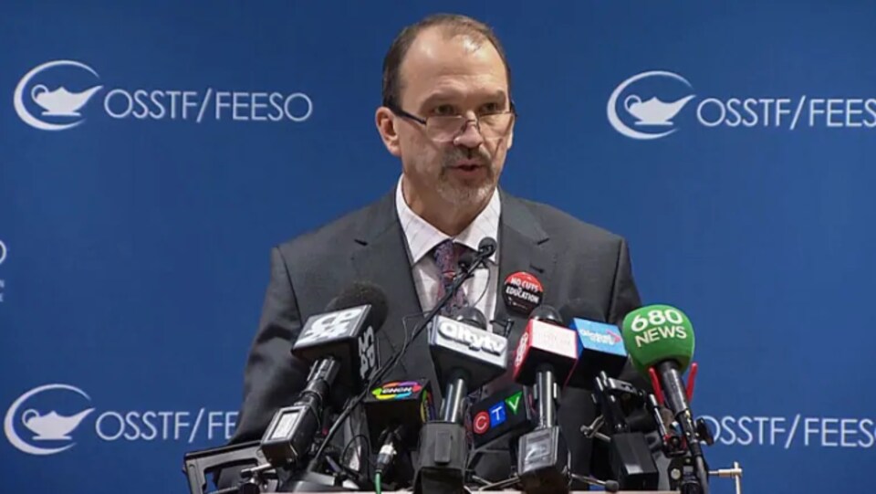 Un homme en conférence de presse avec les micros des grands réseaux posés sur son lutrin.