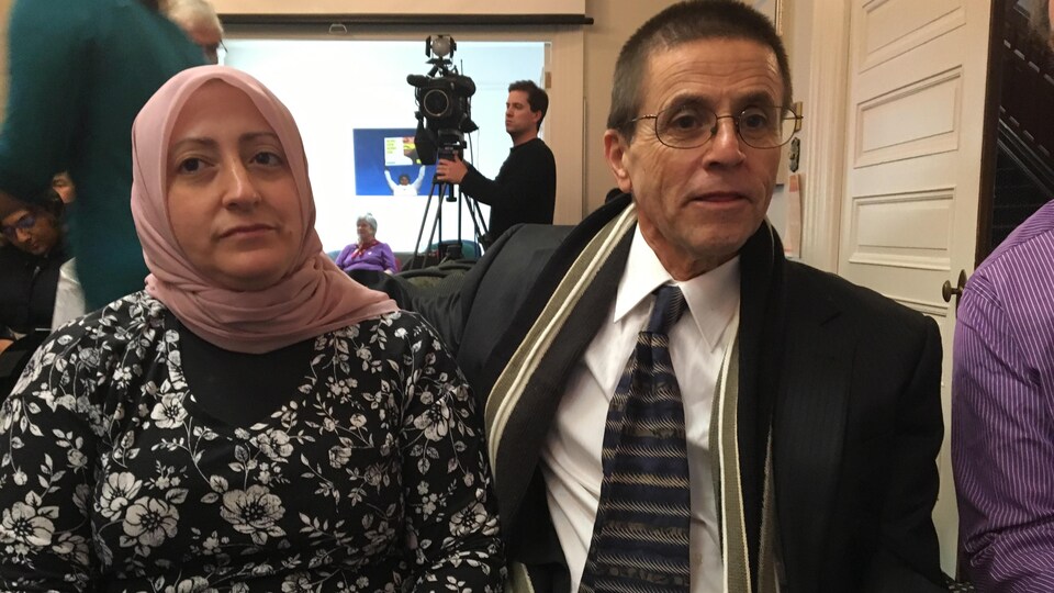 Hassan Diab, assis, tient sa femme par les épaules dans une salle de conférence.