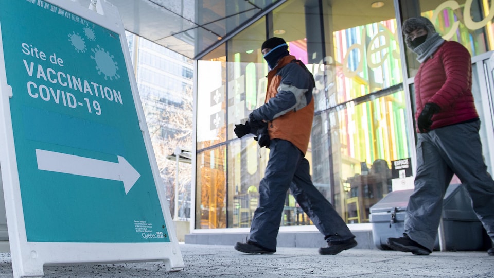 Deux hommes chaudement habillés marchent près d'une pancarte sur laquelle une flèche indique le site de vaccination du Palais des congrès de Montréal.