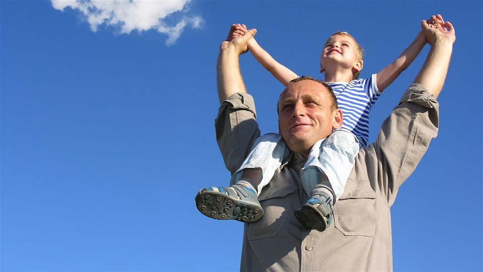 Un père de famille, souriant, avec son jeune garçon sur les épaules.