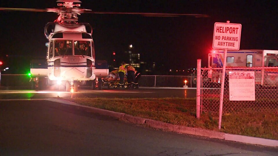 Un hélicoptère se pose en soirée sur la piste d'un héliport. Derrière près de la piste, on voit des ambulanciers debout autour d'une civière.