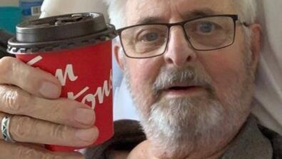 Un homme aux cheveux blancs dans un lit d'hôpital avec une tasse de café.