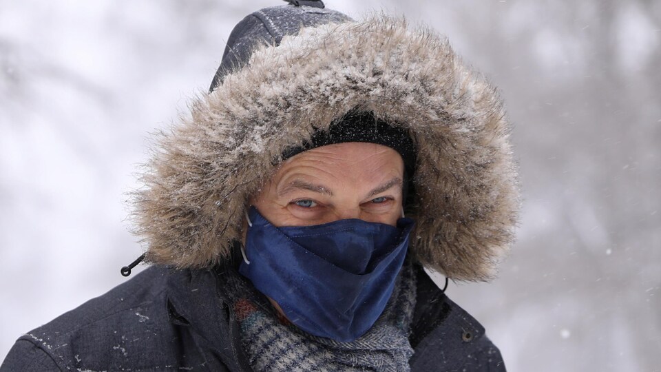 Un homme le visage couvert d'un masque et le capuchon de son manteau d'hiver sur la tête regarde la caméra et plisse les yeux alors qu'il marche à l'extérieur pendant une tempête de neige.
