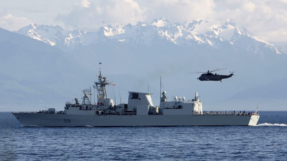 Devant les montagnes de l'État de Washington en toile de fond, un hélicoptère Sea King survole le navire HMCS Vancouver à Esquimalt, en Colombie-Britannique.