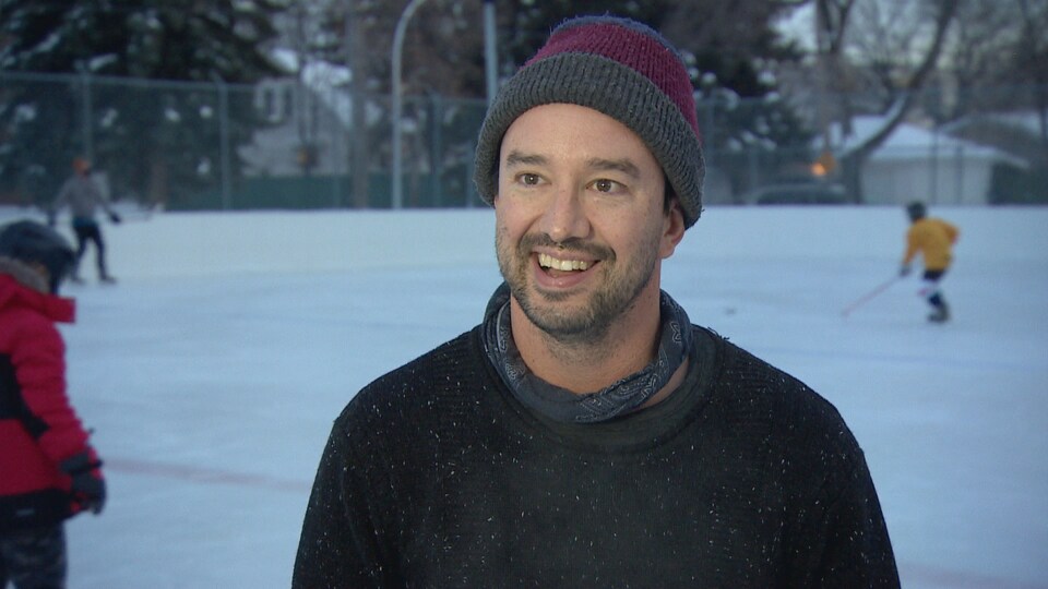L'homme sourit à la caméra sur la patinoire. Des enfants jouent au hockey derrière lui.