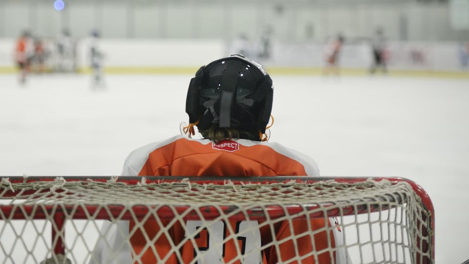 Un enfant est gardien de but de hockey. Il est de dos et fait face au reste des joueurs sur une patinoire intérieure.